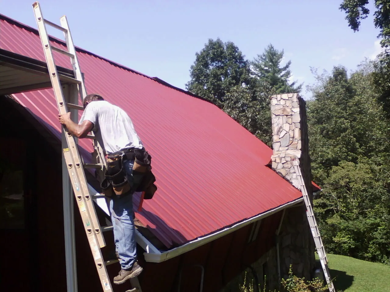 Roofer on a ladder beside a red metal roof and stone chimney — Soffit & Fascia Repair by Roofing Brothers in Danielsville